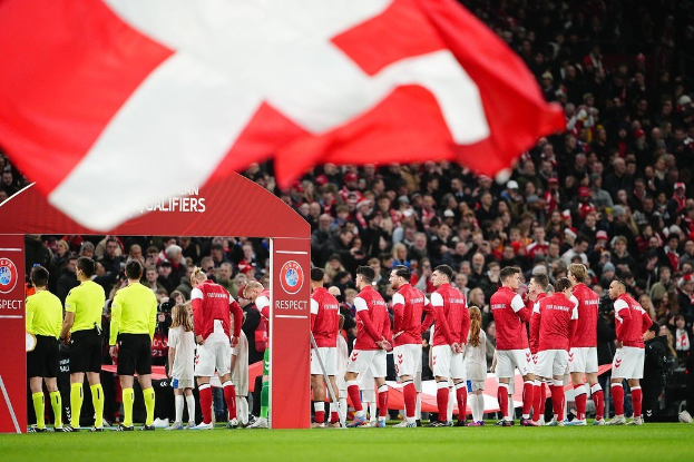 Eine Gruppe von Menschen auf einem Fußballfeld mit einer roten und weißen Fahne im Vordergrund, einem Bogen mit der Aufschrift "Bayern München vs Bayern München Wett-Tipps & Vorschau" im Hintergrund und einer großen Menge im Stadion.