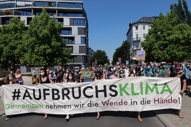 Gruppe von Menschen mit Masken, die ein Transparent mit der Aufschrift "Aufbruchsklima" vor einem Gebäude mit Bäumen und einem klaren Himmel halten.