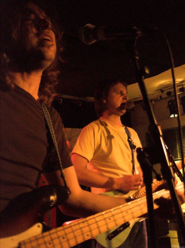 Ein Musikkonzert mit zwei Sängern: Einer in einem weißen Hemd mit Mikrofon, der eine grüne Gitarre hält, und ein anderer in einem schwarzen T-Shirt mit Brille, der eine schwarze Gitarre hält, mit Bühnenbeleuchtung und anderen Instrumenten im Hintergrund.