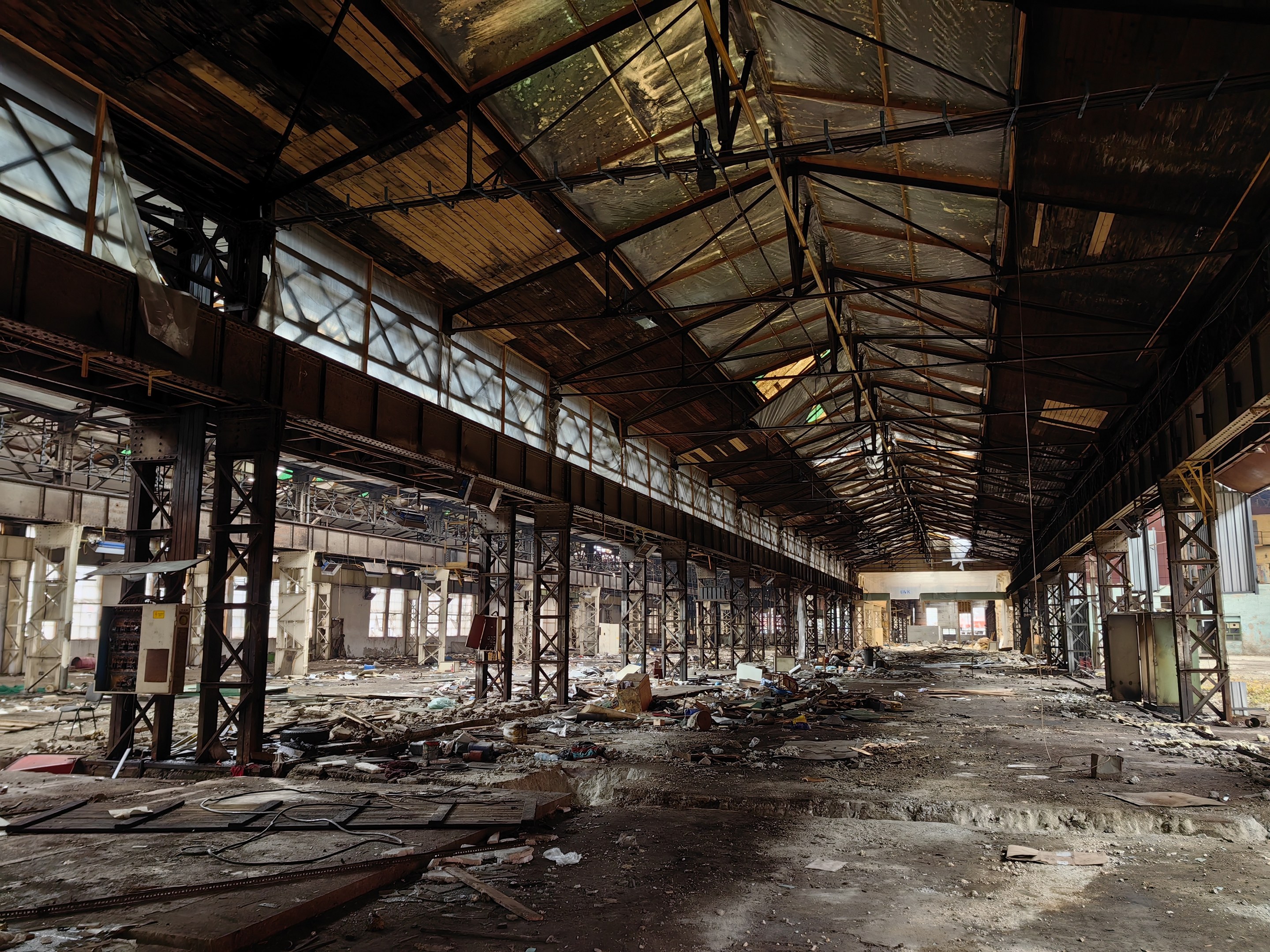 Verlassene Fabrikhalle mit verstreuter Trümmern, kaputten Fenstern, Metallstangen, Deckenleuchten und einem beschädigten Dach.