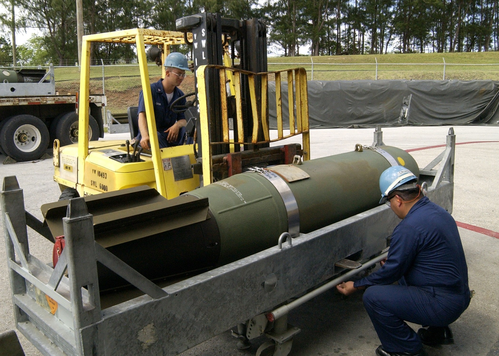 Zwei Männer in blauen Uniformen und Helmen arbeiten an einer großen Metall-Fragmentierungsbombe im Freien mit einem Zaun, Bäumen und einem klaren blauen Himmel im Hintergrund.