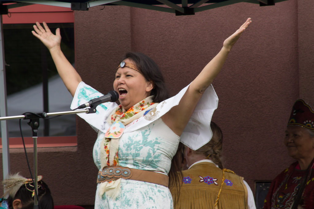 Eine Frau in einem weißen Kleid steht auf einer Bühne, schreiend in ein Mikrofon, mit einer Wand und einem Eisenrahmen im Hintergrund.