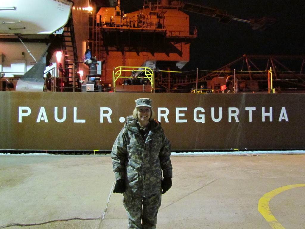 Eine Frau lächelt, trägt eine Mütze und Handschuhe und steht vor einem Schiff mit Text darauf.