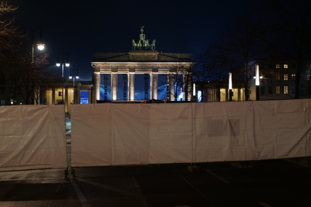 Brandenburger Tor in Berlin nachts, mit einem weißen Tuch bedeckt, von Straßenlaternen beleuchtet, umgeben von Bäumen und Gebäuden mit dem Himmel im Hintergrund.