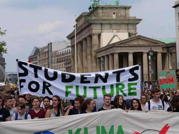 Eine Gruppe von Schülerinnen und Schülern marschiert in Berlin mit einer bunt bemalten "Students for Future"-Fahne vor einer Kulisse aus Gebäuden, Bäumen und Himmel.