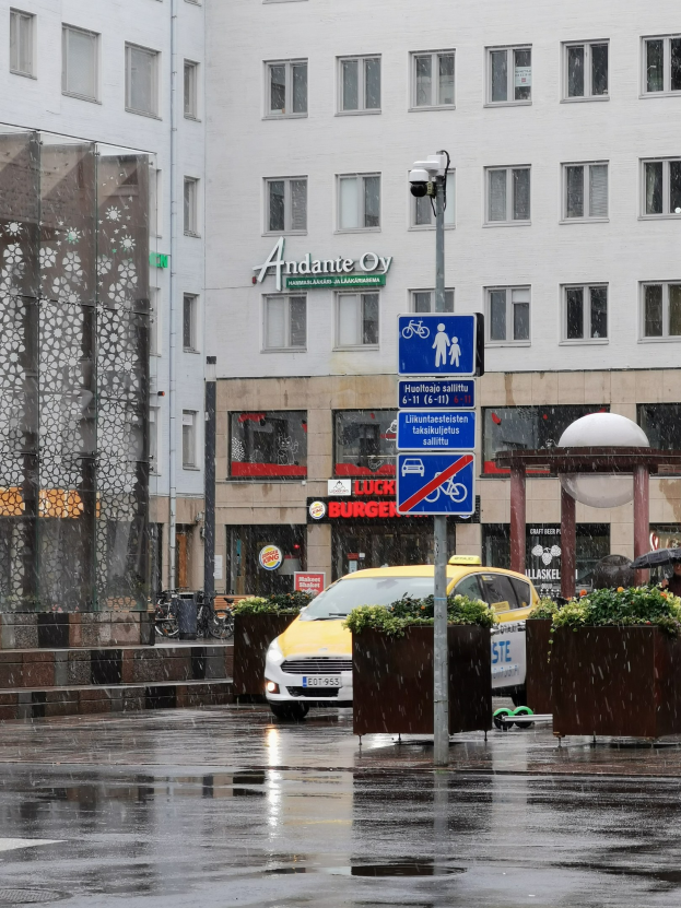 Gelbes Taxi fährt auf regennasser Stadtstraße mit einer Person unter einem Schirm, einem Hinweisschild, Blumenpflanzen und einem Gebäude mit Fenstern im Hintergrund.