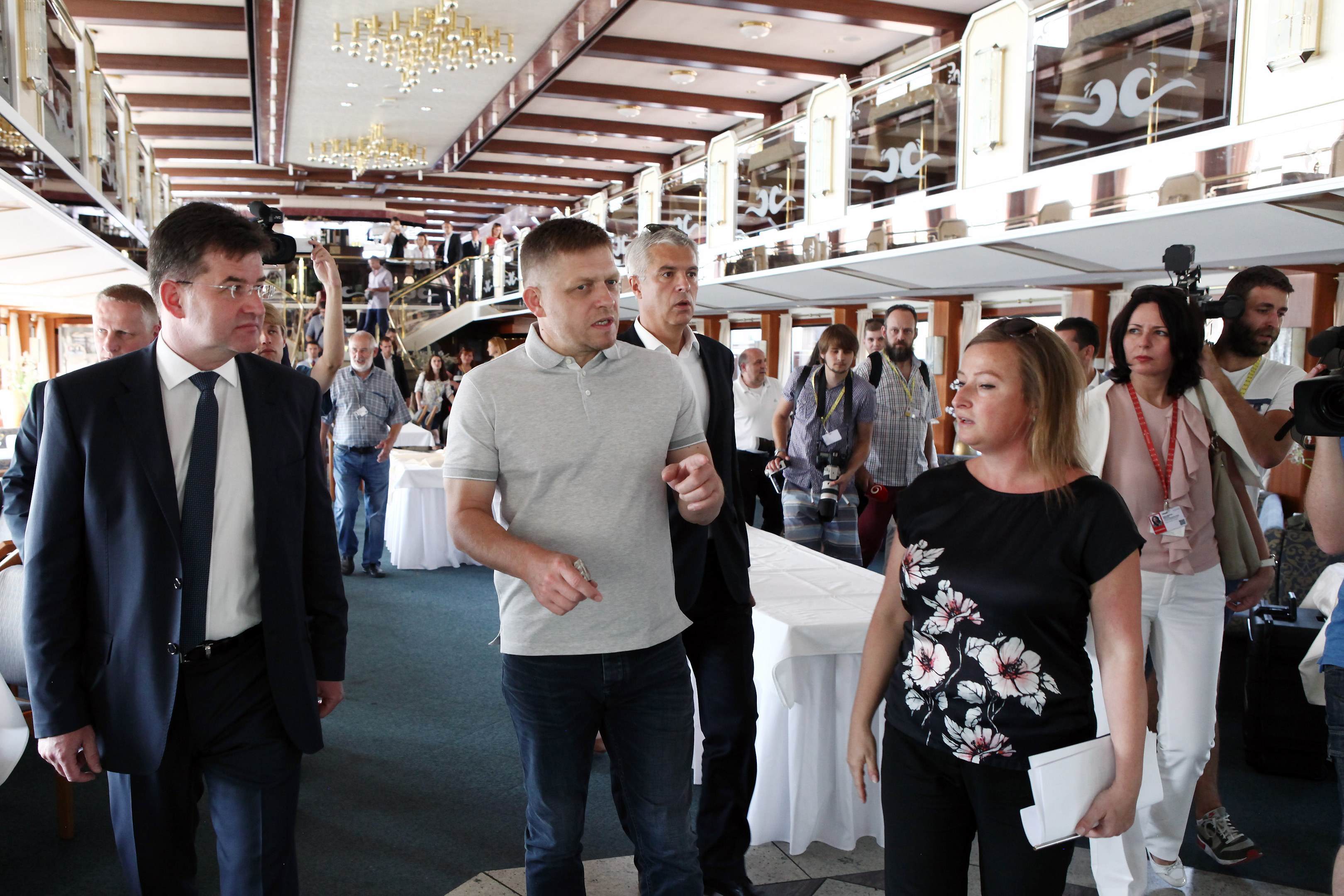 Eine Gruppe von Menschen, die einen Gang auf einem Kreuzfahrtschiff entlanggehen, einige halten Kameras, mit Tischen, Stühlen, Kronleuchtern, Geländern und Glasfenstern im Hintergrund.