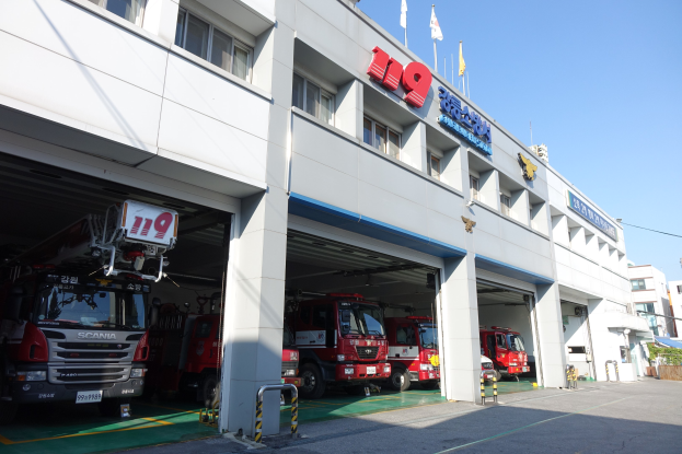 Feuerwache mit mehreren geparkten Feuerwehrautos, einem Gebäude mit Fenstern, Säulen und Namensschildern sowie Flaggen im Vordergrund, vor einer Kulisse anderer Gebäude und einem klaren blauen Himmel.