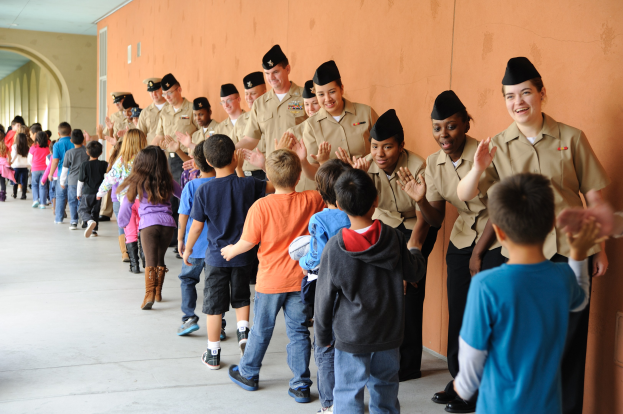 Eine Gruppe von Kindern in Uniformen, die einen Flur mit einer Wand auf der rechten Seite und Deckenleuchten darüber entlanggehen.
