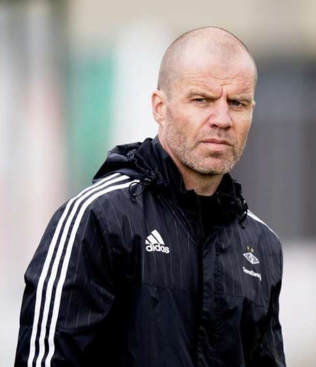 Ein kahlköpfiger Mann in einer schwarzen Jacke steht auf einem Fuballfeld, trägt eine marineblaue Jacke mit weißen Streifen und einem weißen Kragen und hat eine ernste Miene.