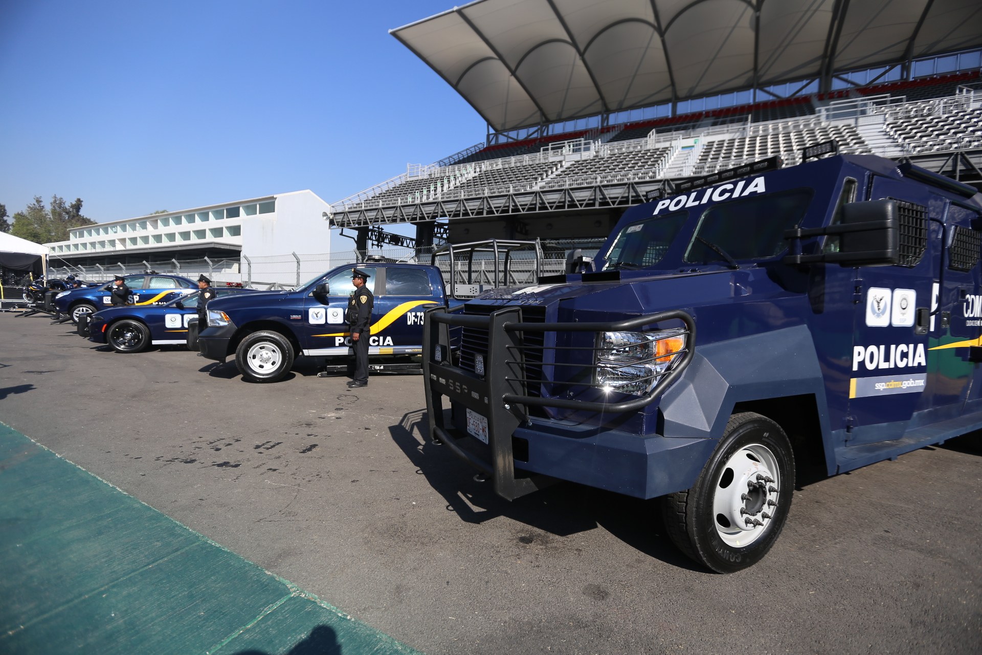 Eine Reihe von Polizeiwagen vor einem Stadion geparkt, mit Menschen auf der Straße im Vordergrund, Gebäuden, Bäumen und einem klaren blauen Himmel im Hintergrund.