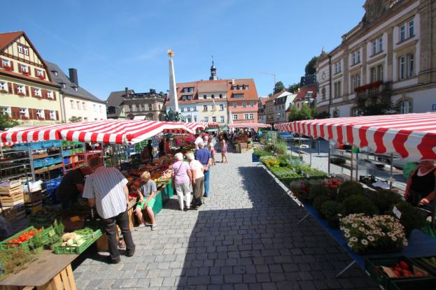 Ein belebter Markt im historischen Zentrum von Heidelberg, Deutschland, mit Menschen, die spazieren gehen, auf Bänken sitzen und um Zelte und Tische mit Körben voller Gemüse sowie Gebäude mit Fenstern im Hintergrund unter einem klaren blauen Himmel.