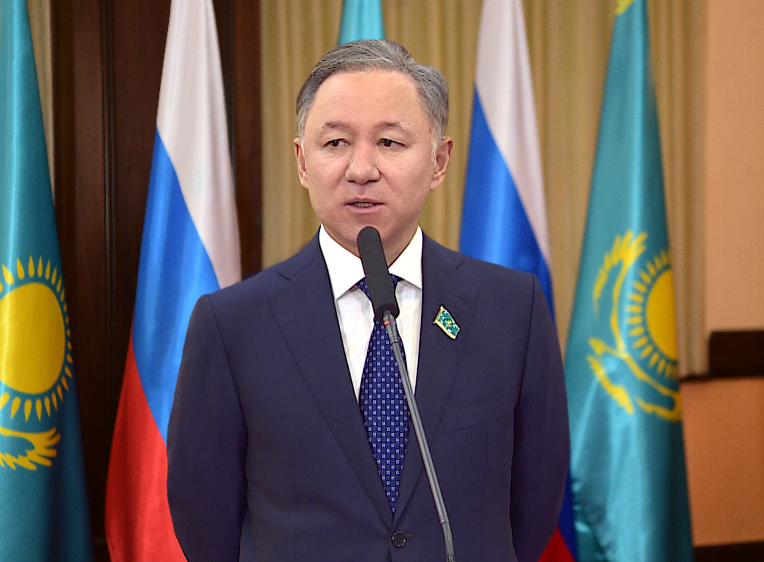 Ein Mann in Anzug und Krawatte spricht an einem Podium mit Mikrofonen, umgeben von Flaggen und einer Wand im Hintergrund.