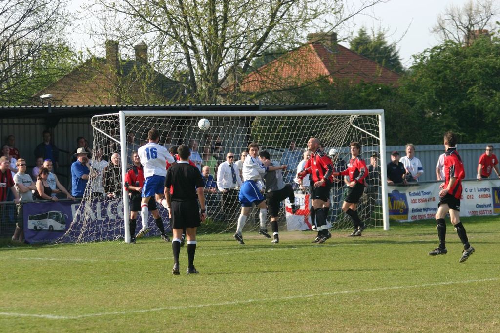 Fussballspieler sind in einem Spiel auf einem Feld mit einem Tornetz beteiligt, während Zuschauer dahinter stehen, mit Bäumen und Häusern im Hintergrund.
