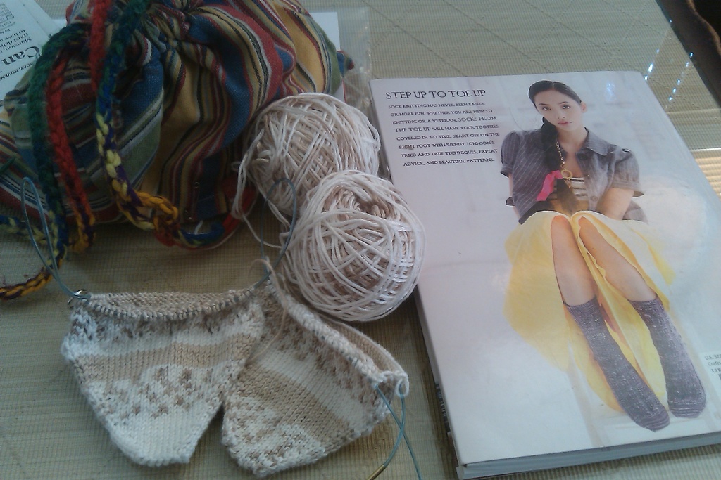 Ein Buch, eine Wolle, ein Stück Stoff und einige Papiere sind auf einem Tisch angeordnet.