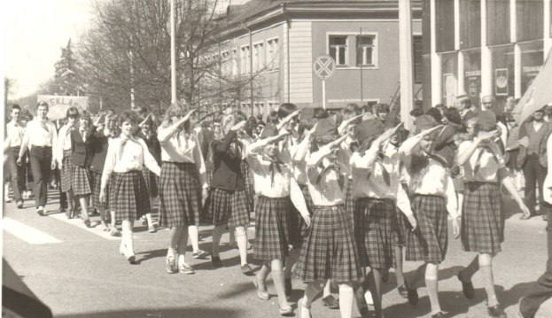 Eine Gruppe von Menschen in Schuluniformen, die eine Straße entlang marschieren, einige halten Fahnen, mit Bäumen, Polen, Schildern, Gebäuden mit Fenstern und einem klaren Himmel im Hintergrund, dargestellt in Schwarz-Weiß.