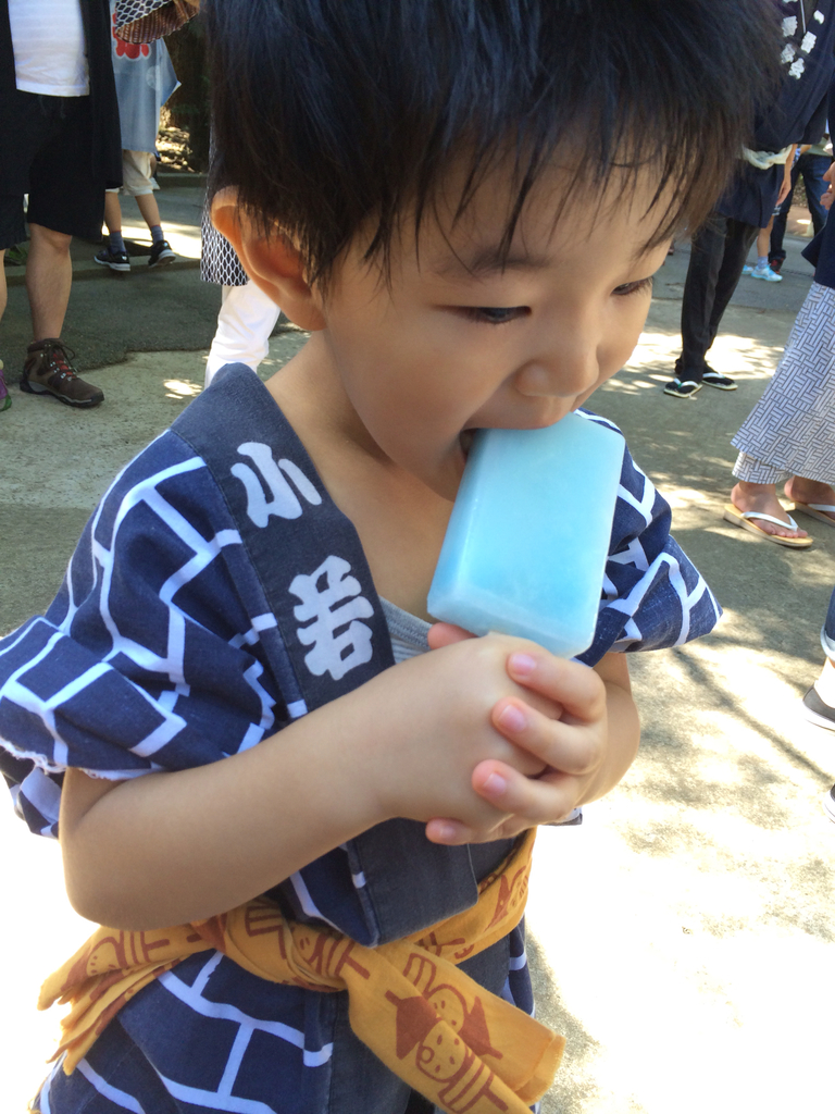Ein Kind in einem blauen Outfit leckt ein Eis, während Menschen im Hintergrund stehen.