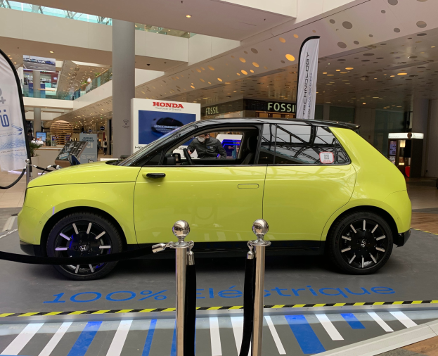 Gelber wasserstoffbetriebenes Elektroauto vor einem Gebäude mit Absperrpfosten, Bannern, Menschen, Schildern, einer Topfpflanze, Deckenleuchten und Lüftern.