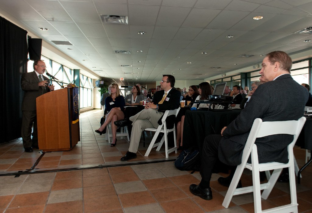 Menschen, die in Stühlen sitzen und einem Mann zuhören, der in der Nähe eines Podiums steht und in ein Mikrofon spricht, mit Fenstern im Hintergrund.