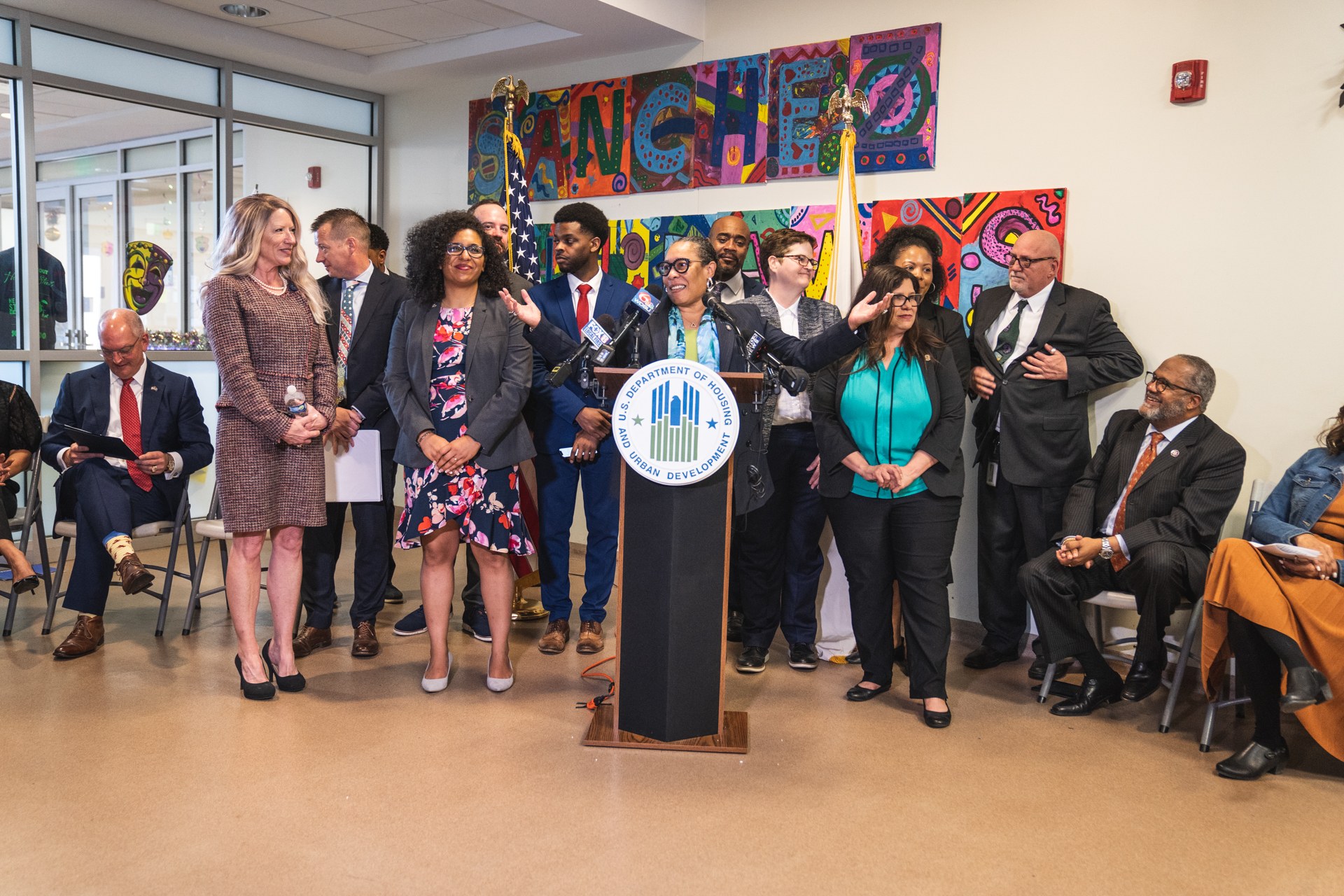 Gruppe von Menschen vor einem Rednerpult mit einem Mikrofon und einer Tafel mit der Aufschrift "Sancho-Stadtrat tritt dem Stadtrat bei", mit sitzenden und stehenden Zuschauern, Fahnen, Feuerlöscher und Deckenleuchten im Hintergrund.