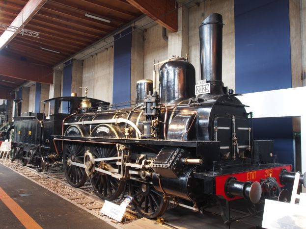 Eine große, alte Dampflokomotive in einem Museums-Setting, umgeben von Eisenbahnschienen, Informations-Tafeln und anderen Ausstellungen, mit einer Wand und Deckenbeleuchtung im Hintergrund.