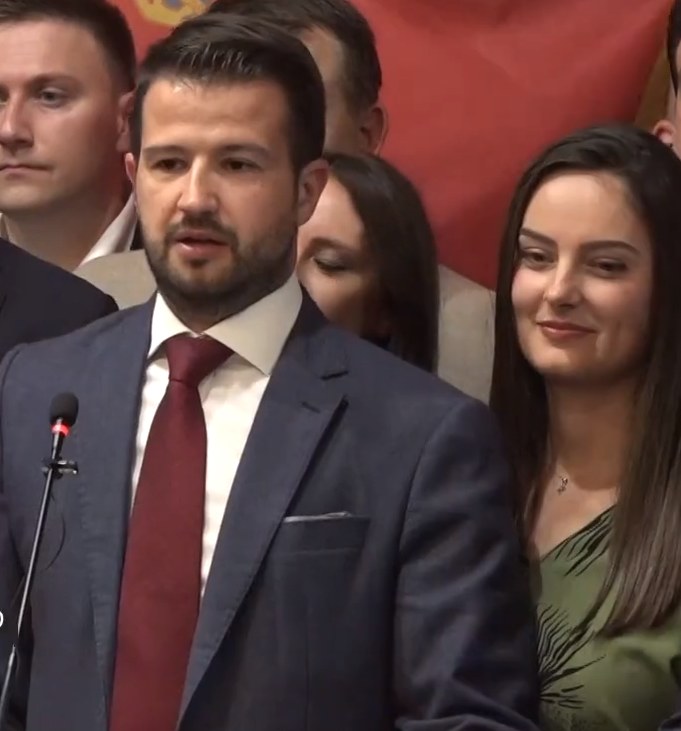 Mann in Anzug und Krawatte, der an einem Rednerpult spricht, vor einer Menge mit einem Banner im Hintergrund.