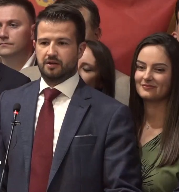 Mann in Anzug und Krawatte, der an einem Rednerpult spricht, vor einer Menge mit einem Banner im Hintergrund.