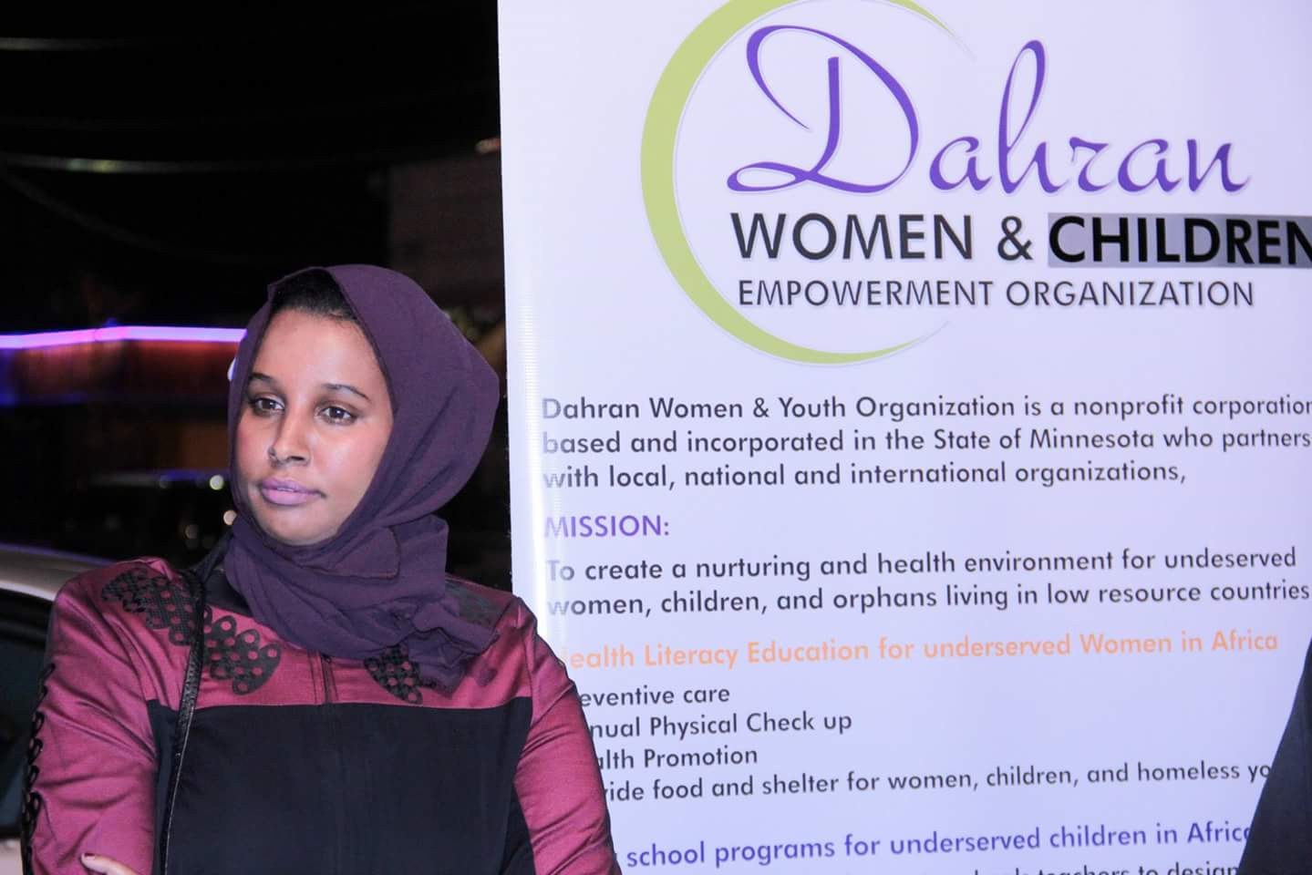 Eine Frau in einem weißen Hemd und blauen Jeans steht entschlossen vor einem Banner mit der Aufschrift "Dahran Women & Children Empowerment Organization", wobei ein Auto im Hintergrund zu sehen ist.