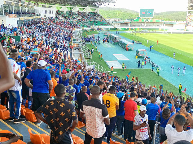 Menge Menschen vor einem eingezäunten Fußballfeld mit Hügeln und einem klaren blauen Himmel im Hintergrund während der Afrikameisterschaft 2019.