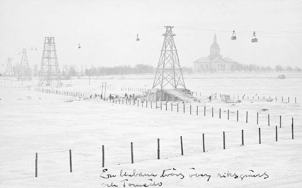 Schwarze-Weiß-Foto eines Skilifts in einer verschneiten Wiese mit Stützpfählen und Drähten, umgeben von Bäumen und einem Gebäude im Hintergrund, mit Text unten.