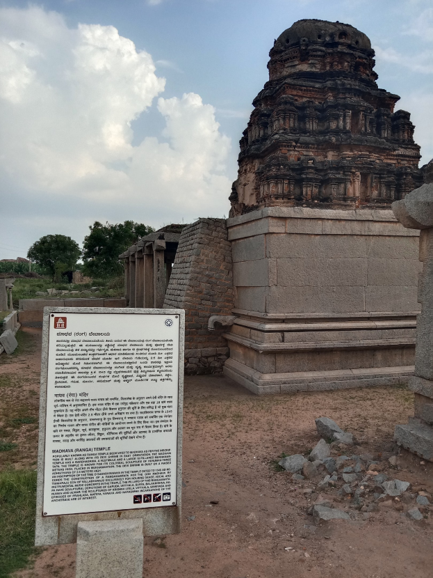Ein Schild vor einem Tempel in einer grasbewachsenen Wiese mit verstreuten Steinen und Bäumen, unter einem bewölkten Himmel, das 'Madhya Pradesh - Madhya Pradesh Temple' lautet.