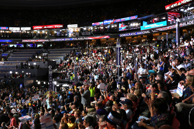 Große Menschenmenge auf der Democratic National Convention 2016 in Philadelphia mit Schildern, Treppe zum hell erleuchteten Podium und Texttafeln drumherum.