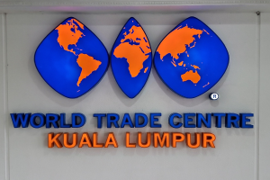 Das Logo des World Trade Centre in Kuala Lumpur, Malaysia, auf einer Wand mit fetter schwarzer Schrift und einem weißen Rand vor einem hellblauen Himmel mit weißen Wolken.