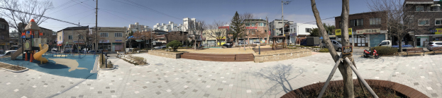 Stadtplatz mit einem zentralen Spielplatz, umgeben von Bäumen, Gebäuden, Strommasten mit Drähten, Fahrzeugen auf der Straße, Informationsschildern und einem sichtbaren Himmel.