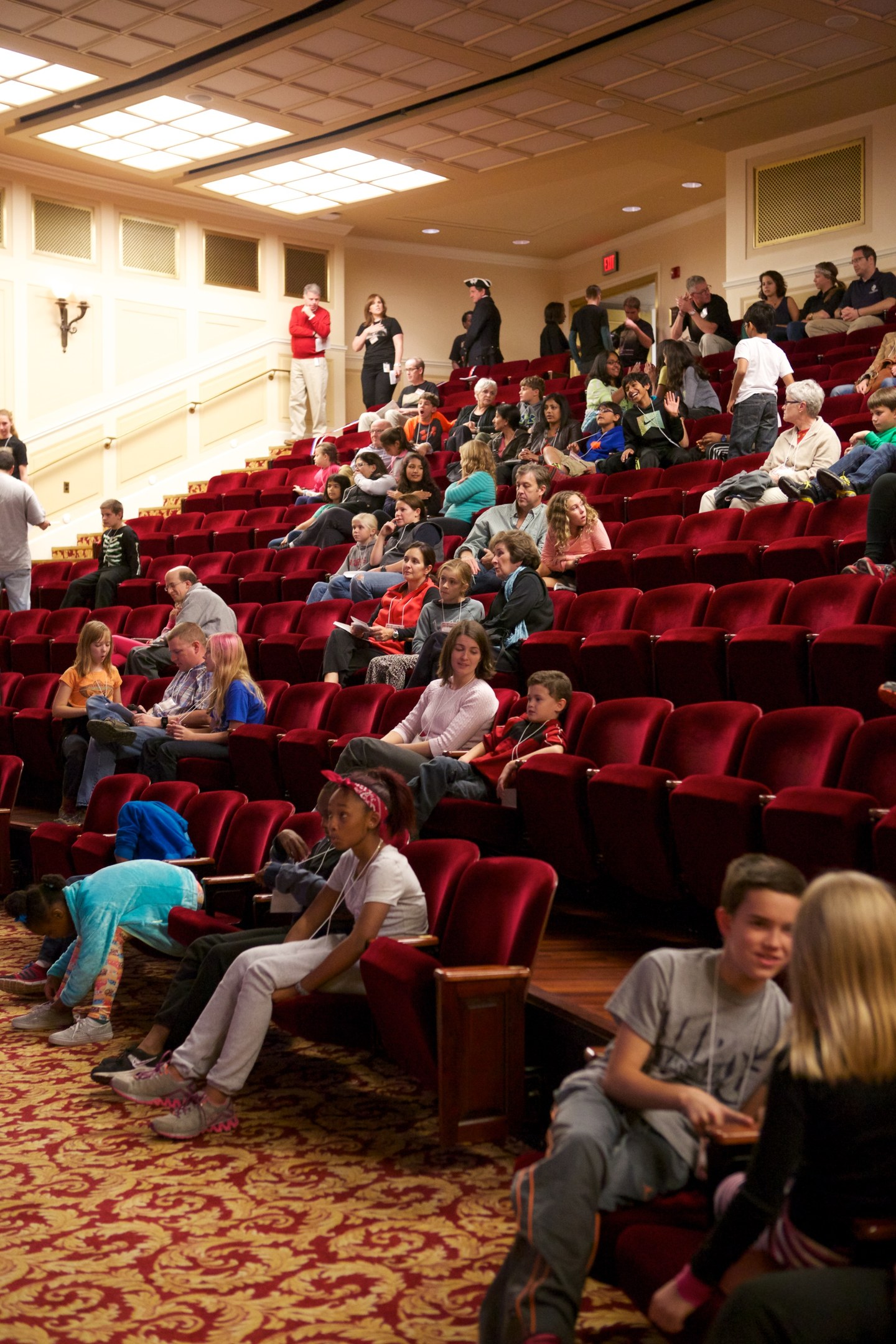 Eine große Gruppe von Menschen, darunter Kinder, die auf Stühlen sitzen und stehen, mit verstreuten Stühlen, Wandlampen und Deckenlampen.
