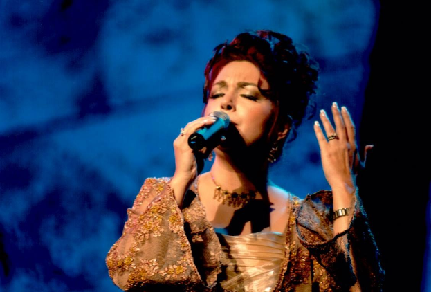 Frau in goldnem Kleid, singend ins Mikrofon auf der ESC-Bühne mit blau-schwarzem Hintergrund.