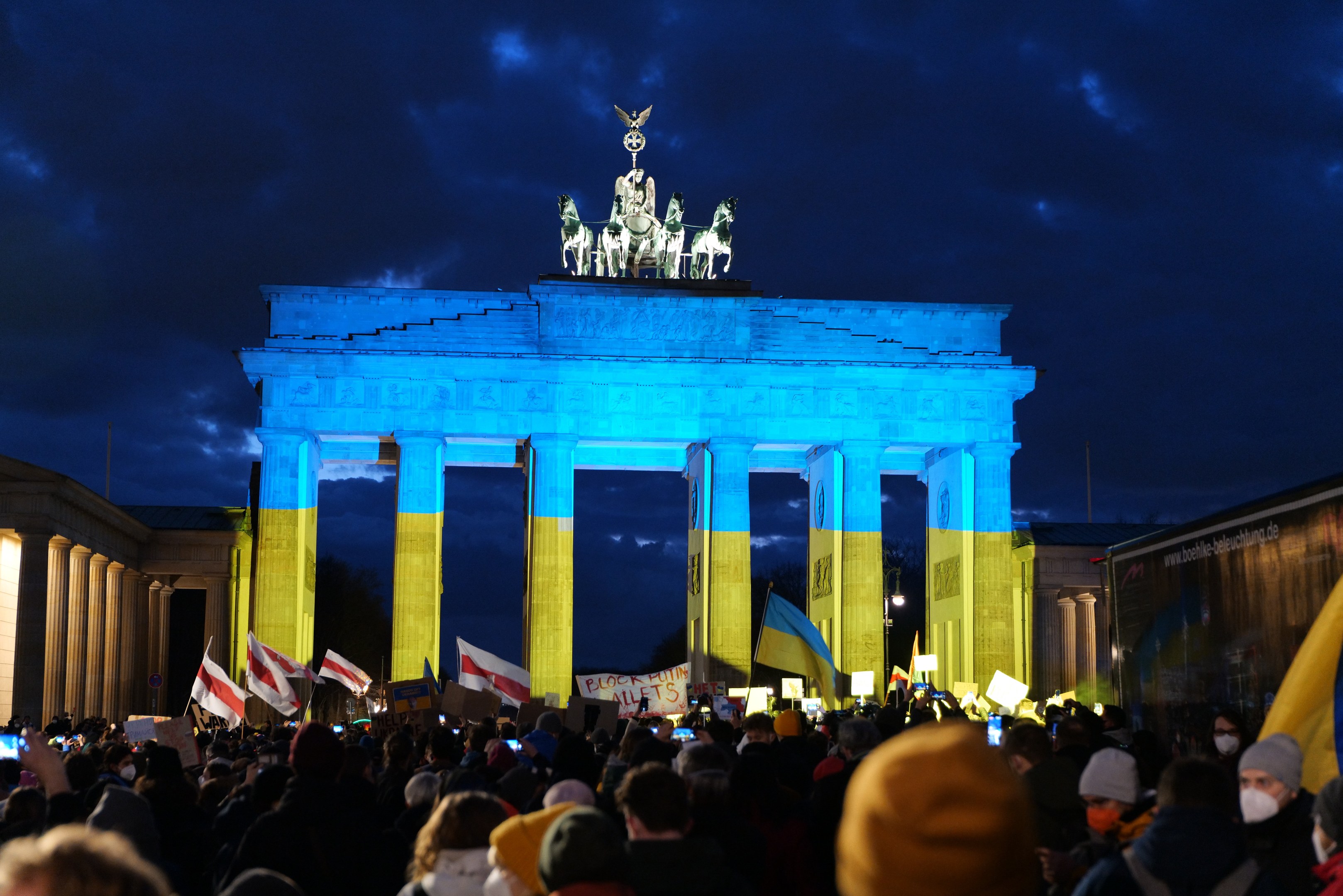 Eine Menschenmenge mit Fahnen und Schildern steht vor dem Brandenburger Tor in Berlin, Deutschland, mit einer Fahne auf der rechten Seite und bewölktem Himmel.