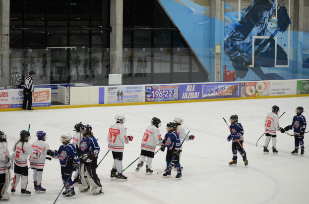 Gruppe junger Menschen beim Eishockey auf einer Indoor-Eisfläche, mit Helmen und Schlägern, einer Person auf der linken Seite und Bannern, Säulen und Glaswänden im Hintergrund.