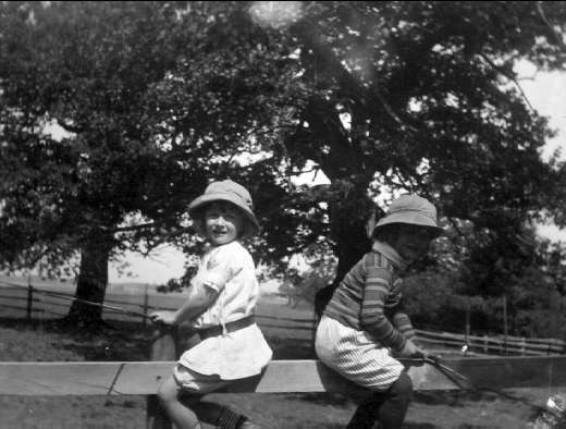 Zwei junge Jungs in Mützen halten Baseballschläger in einem grünen Park mit Bäumen und einem Zaun im Hintergrund, in Schwarz-Weiß.