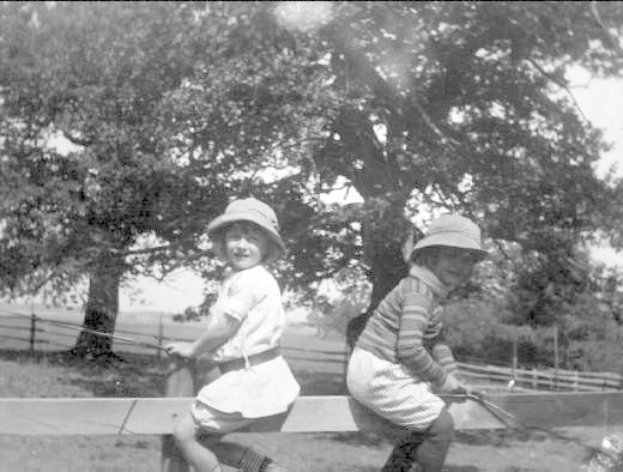 Zwei junge Jungs in Mützen halten Baseballschläger in einem grünen Park mit Bäumen und einem Zaun im Hintergrund, in Schwarz-Weiß.