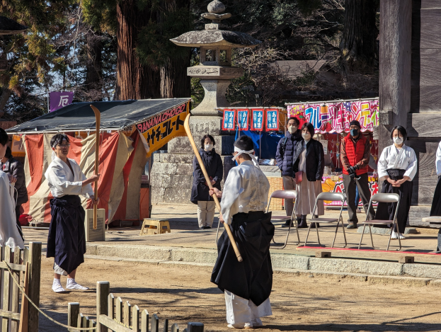 Eine Gruppe von Menschen in traditioneller Kleidung versammelt sich im Freien in Kyoto, einige tragen Masken und halten Holzstöcke, während Stühle, Banner, ein Zelt und ein hölzerner Zaun unter einem klaren blauen Himmel zu sehen sind.