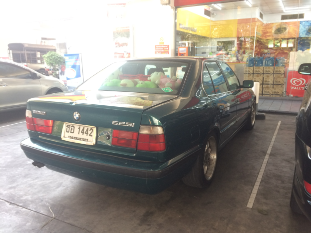 Ein grünes Auto, das auf einem Parkplatz mit einem Teddybär auf dem Rücksitz geparkt ist, neben anderen Fahrzeugen, mit einem Geschäft mit Plakaten und Deckenlampen im Hintergrund.