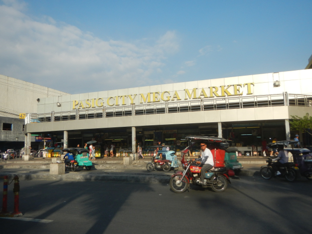 Ein belebter Pasig City Mega Market mit Fahrzeugen auf der Straße, Menschen, die herumlaufen, einem Gebäude mit Text im Hintergrund und einem bewölkten Himmel.