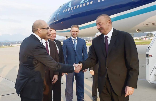 Aserbaidschanischer Präsident İlham Əliyev und türkischer Präsident Recep Tayyip Erdoğan geben sich die Hand vor einem Flugzeug, umgeben von einer Gruppe von Menschen auf dem Rollfeld mit einem Fahrzeug, Gebäuden, Bäumen, Hügeln und einem klaren blauen Himmel im Hintergrund.