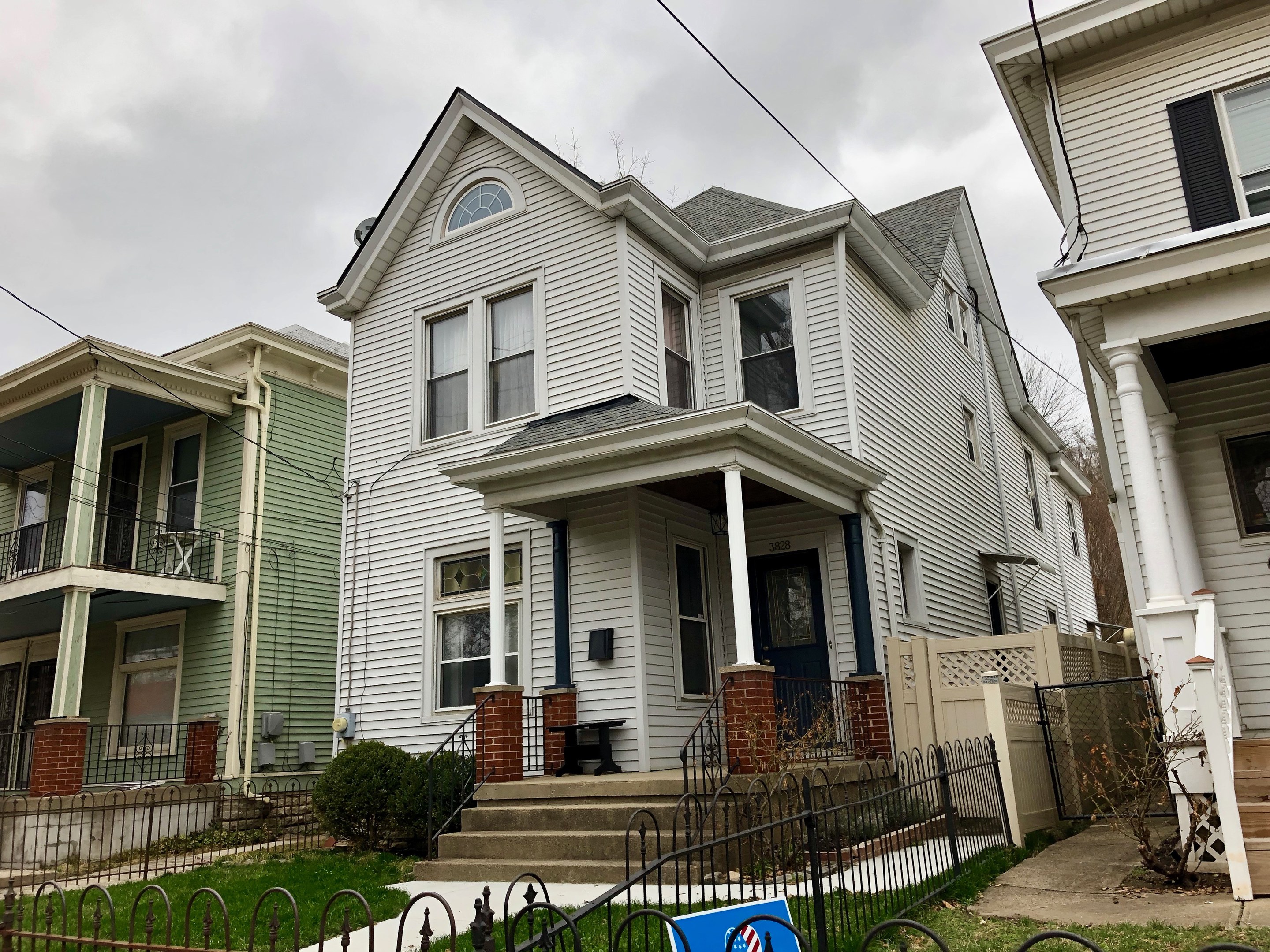 Reihe von Häusern mit Fenstern, Türen, Säulen, Geländern, Stufen und Dächern, umgeben von einem Metallzaun, Gras, Pflanzen, Bäumen und einem bewölkten Himmel, mit einem Mietschild am Boden, das die Adresse und Listen Details anzeigt.