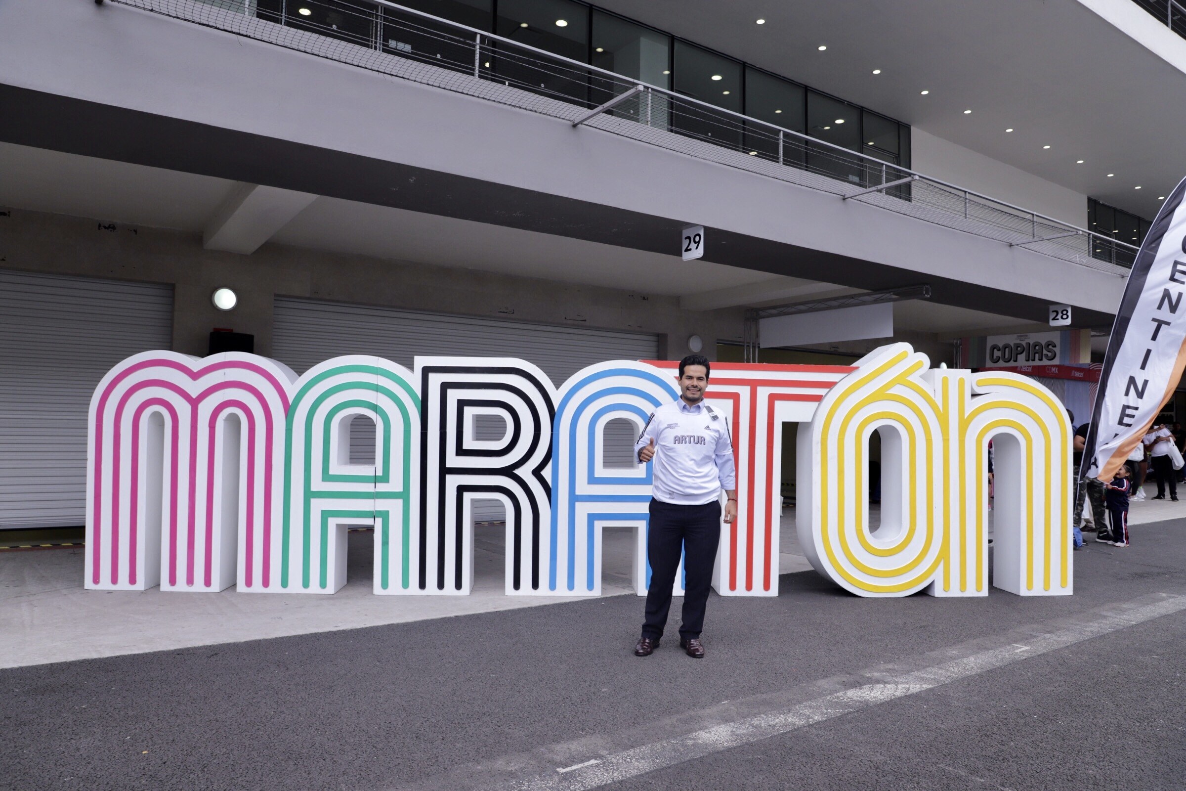 Ein Mann lächelt vor einem "Marathon"-Schild, umgeben von Menschen mit Schildern, mit einem beleuchteten Gebäude im Hintergrund.