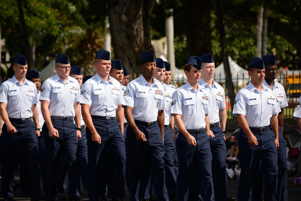 Männer in Uniform auf einer Straße mit einem Eisenzaun dahinter, Bäume und Pfähle im Hintergrund.