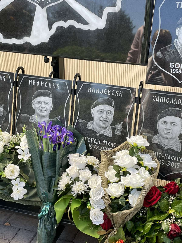 Denkmal mit einem farbenfrohen Blumenstrauß auf einem Gehweg neben einem Schild und Gedenktafeln an der Wand.