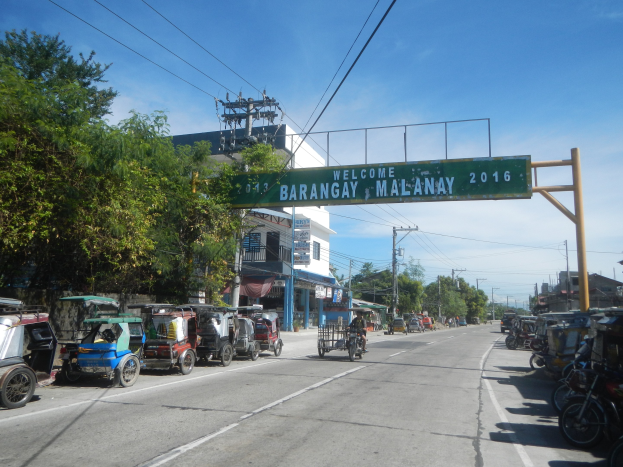 Eine belebte Straße in Barangay, Malaysia, gesäumt von parkenden Rikschas und Fahrzeugen, flankiert von Gebäuden, Bäumen und Strommasten.