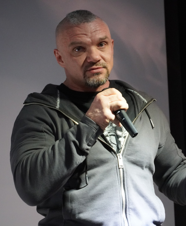 Ein Mann in einer schwarzen Kapuzenjacke und einem schwarzen Anzug mit einem weißen Hemd steht selbstbewusst vor einer Wand und hält ein Mikrofon in der Hand.